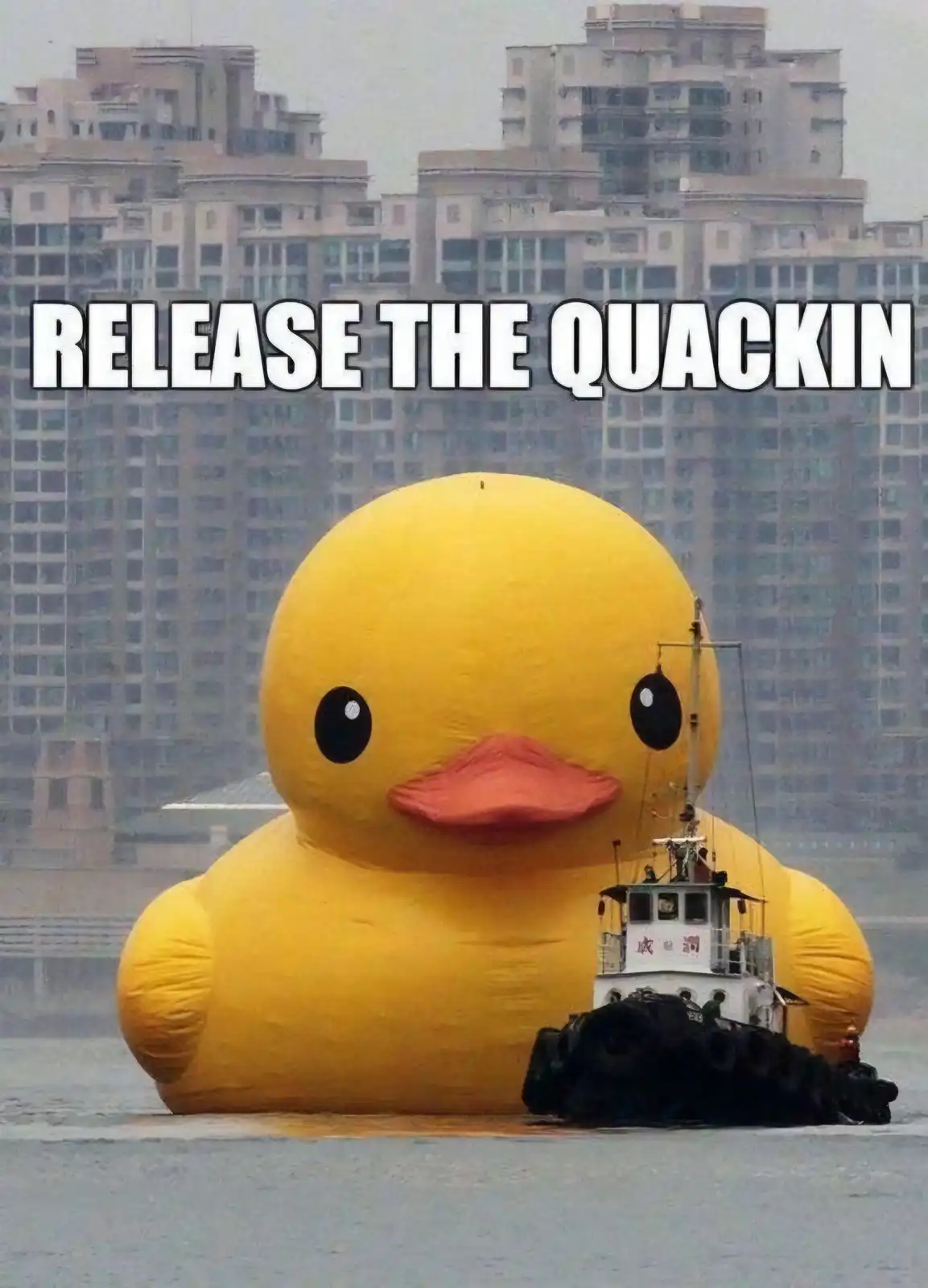 A giant yellow inflatable rubber duck floats in water with a small tugboat beside it, set against a backdrop of dense urban…