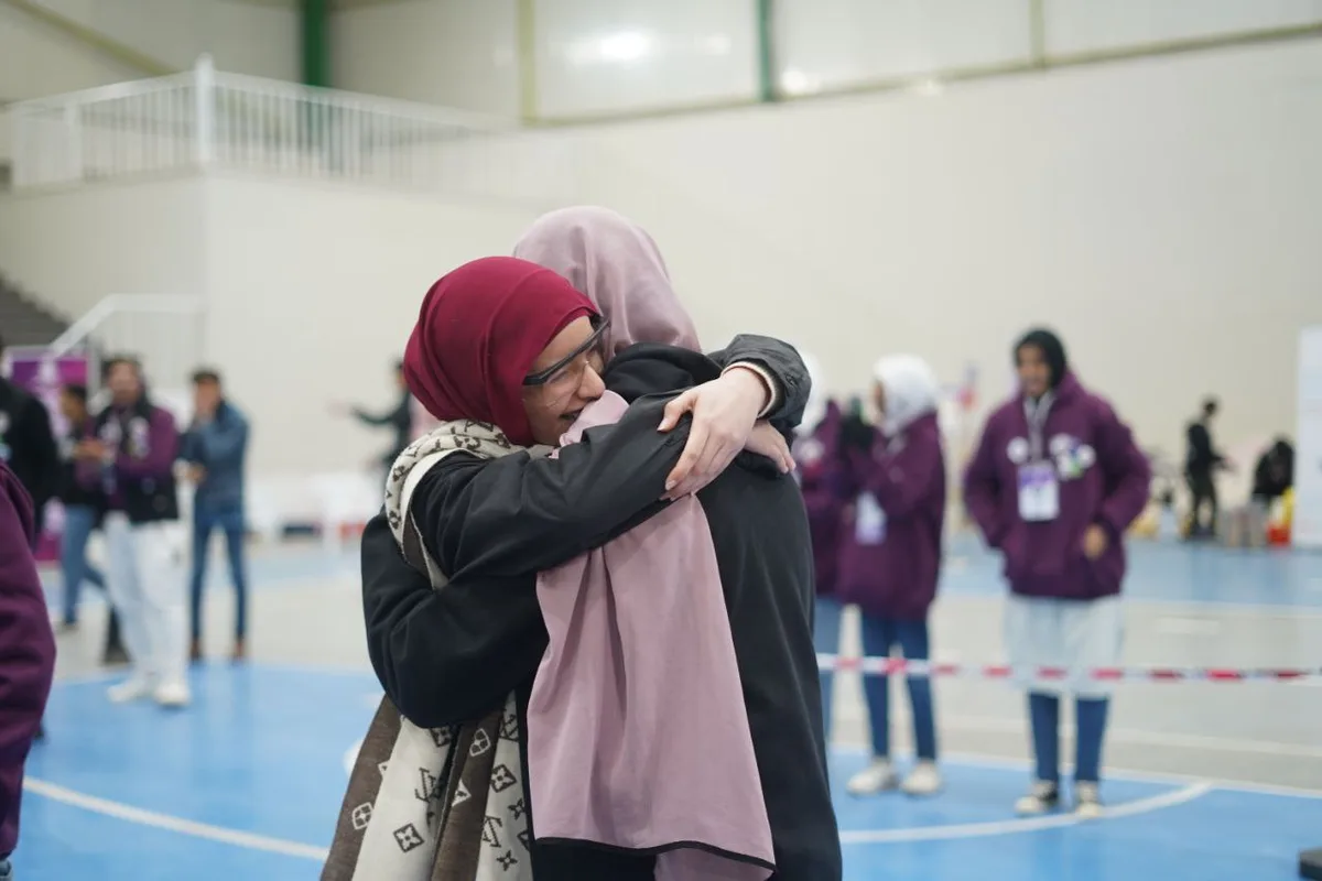 Two women wearing hijabs are hugging tightly in an indoor sports hall with a blue floor. One woman wears a red hijab and…