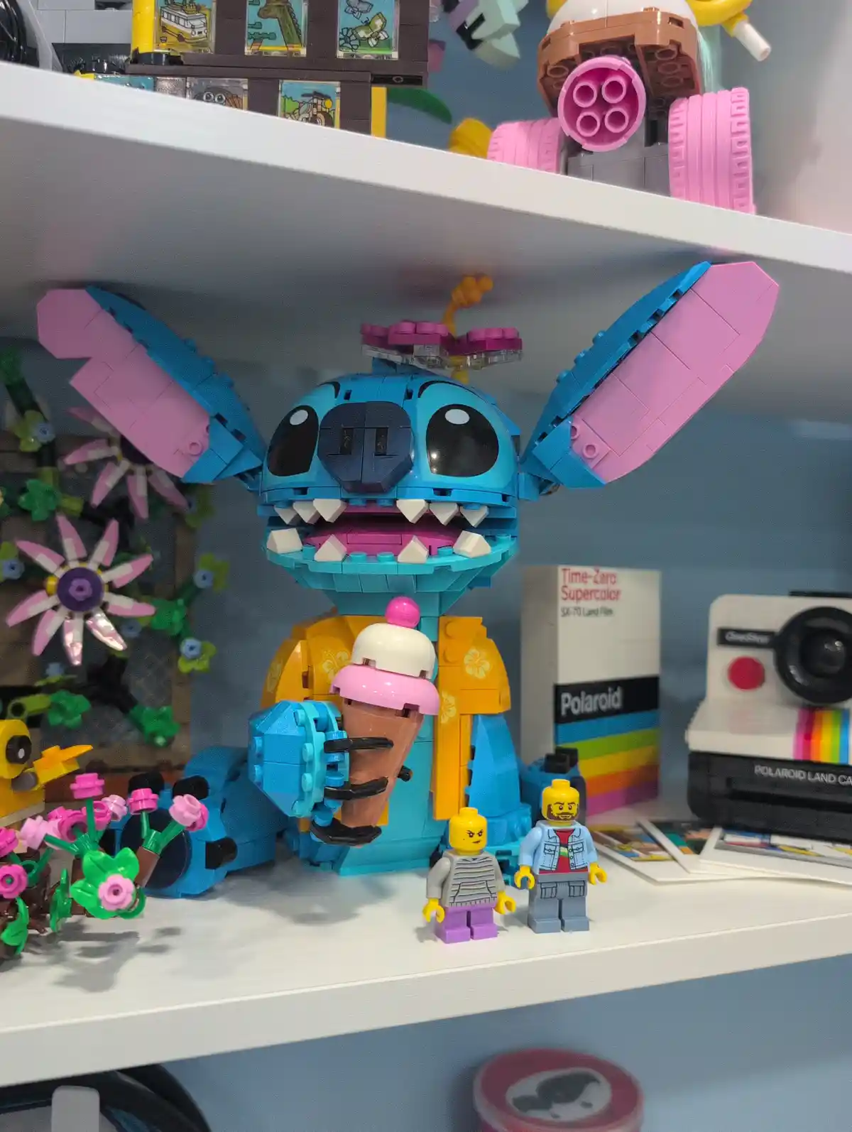 A large blue LEGO Stitch figure with pink ears and a yellow shirt sits on a shelf holding an ice cream cone, surrounded by…