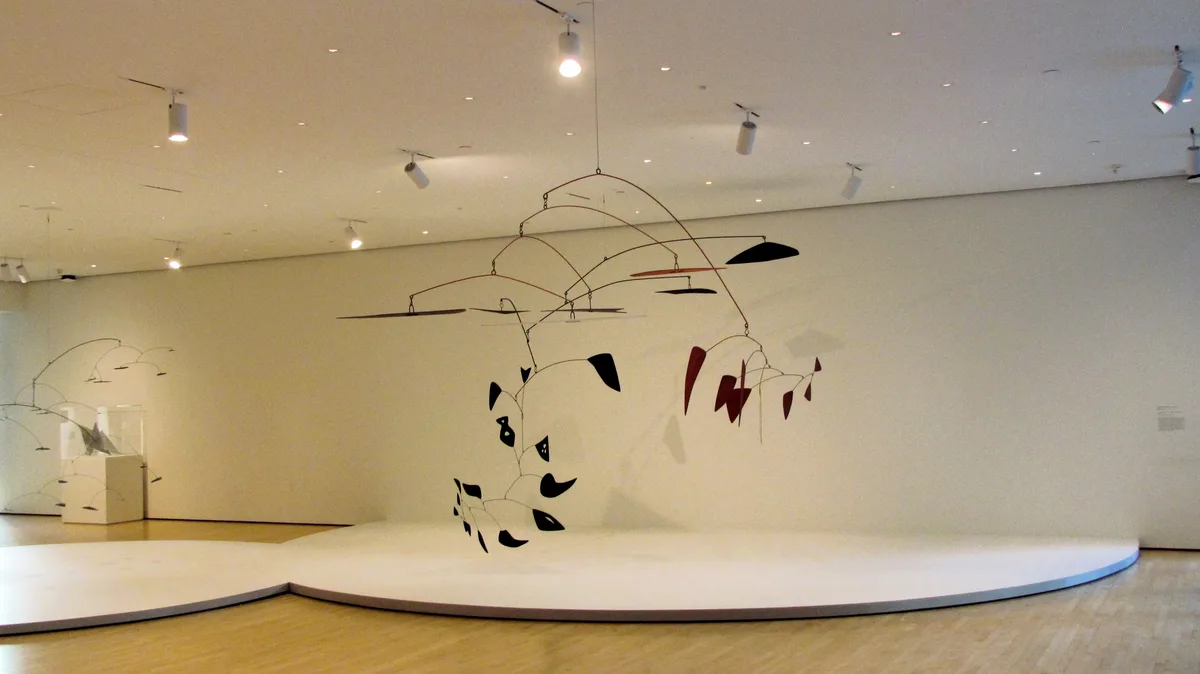 A large, intricate mobile sculpture with black and red geometric shapes suspended from thin rods hangs in a minimalist white…