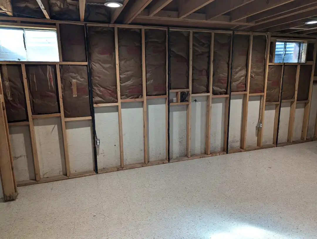 An empty room under construction with exposed wooden wall framing, brown insulation, and a speckled floor. The overlaid text…