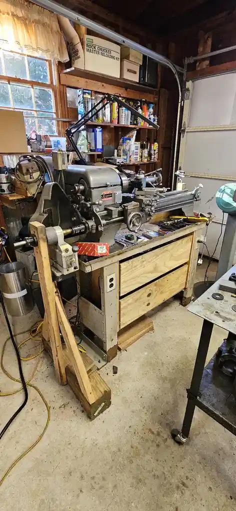 A cluttered garage workshop features a large, vintage Logan lathe machine mounted on a wooden workbench with drawers. The…