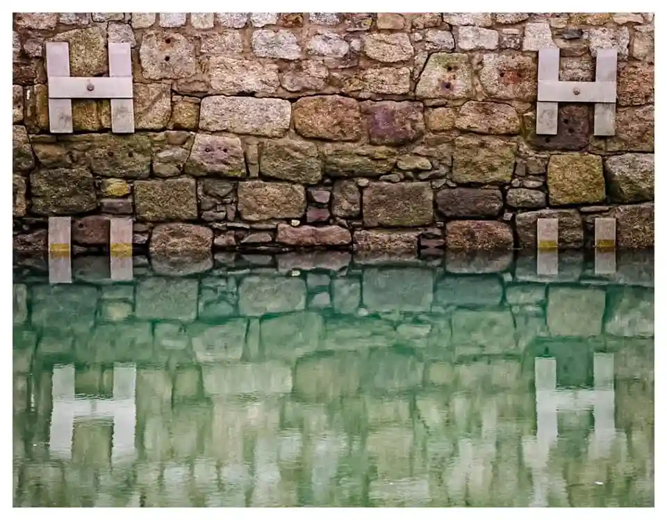 A stone wall with two metal 'H' brackets mounted on it, reflected in calm greenish water below, creating a symmetrical…
