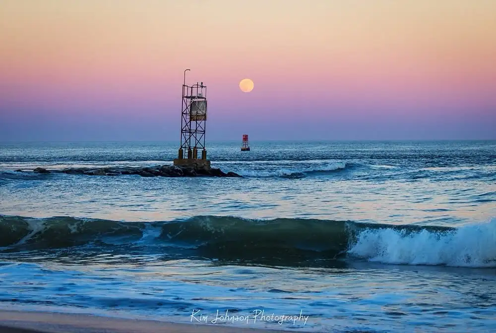 A serene beach scene at dusk or dawn with a full moon visible in the sky, a small lighthouse on rocks in the middle ground…
