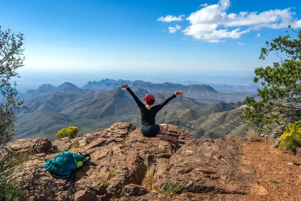 A person with red hair sits on a rocky mountain peak with arms outstretched, facing a vast mountain range under a blue sky…