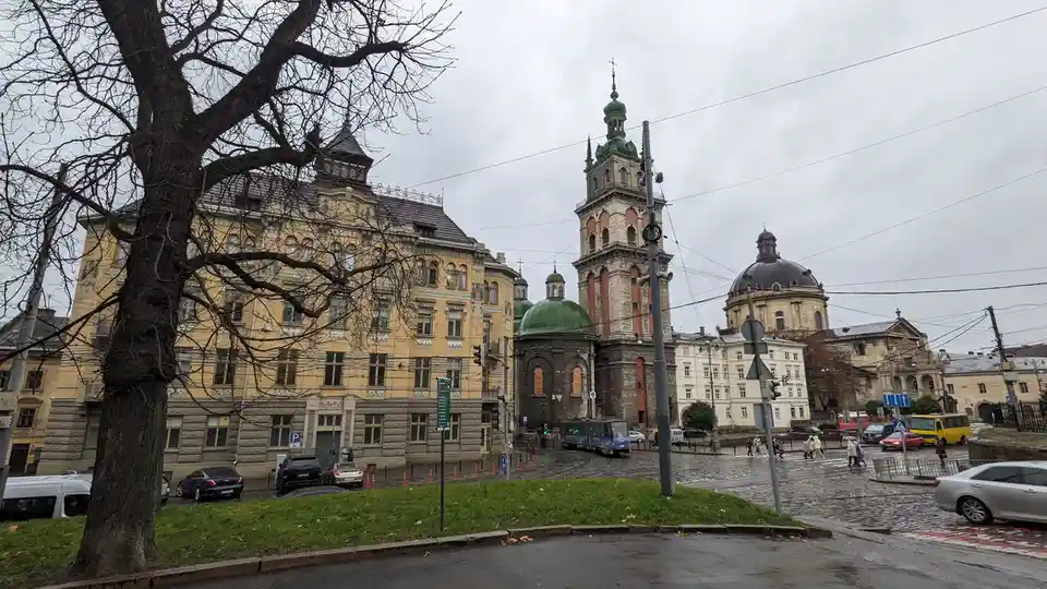 A photo of a European city square with historic buildings, including a church with a tall tower and green domes, under an…