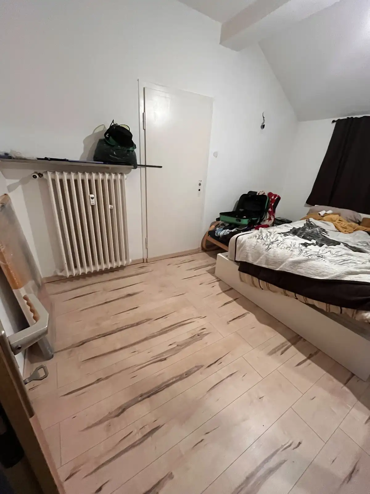 A messy bedroom with a bed covered in clothes and an open suitcase, a radiator with bags on top, and a chair with a plastic…