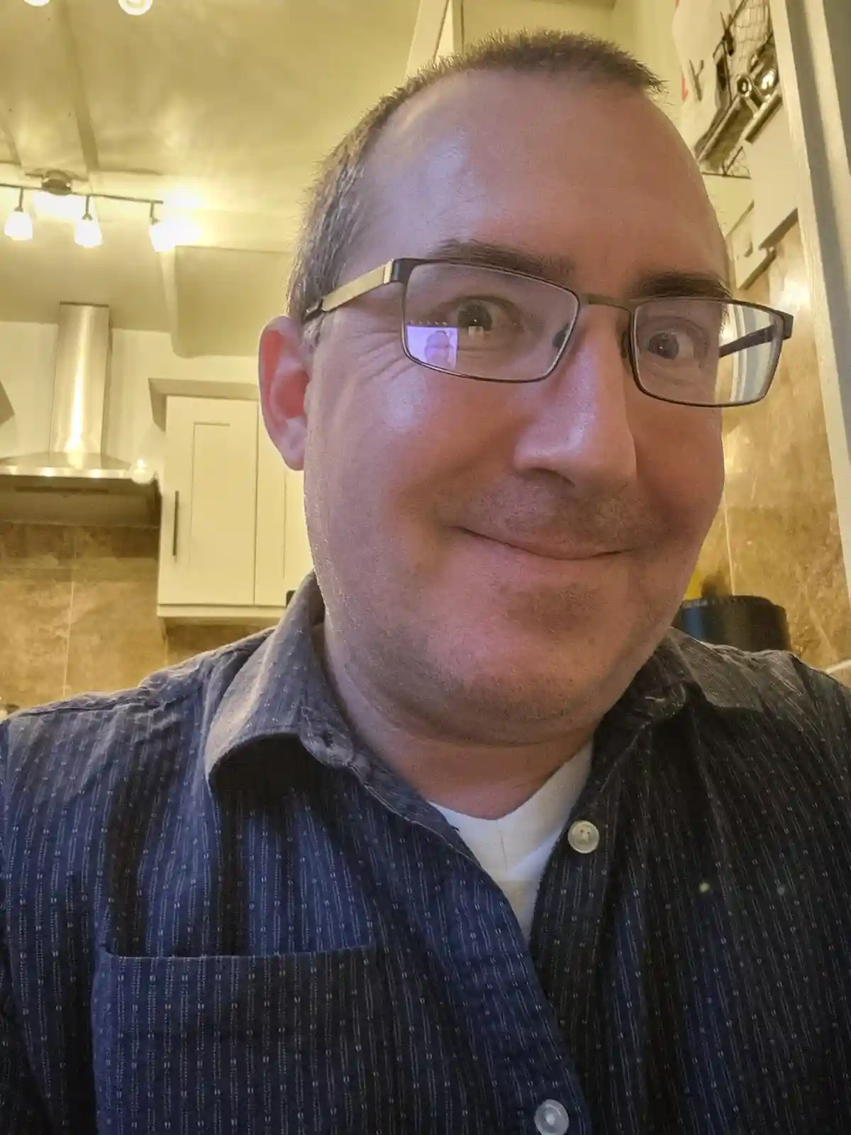 A man with glasses and a slight smirk looks at the camera in a kitchen, with a faint reflection of a video call visible in…