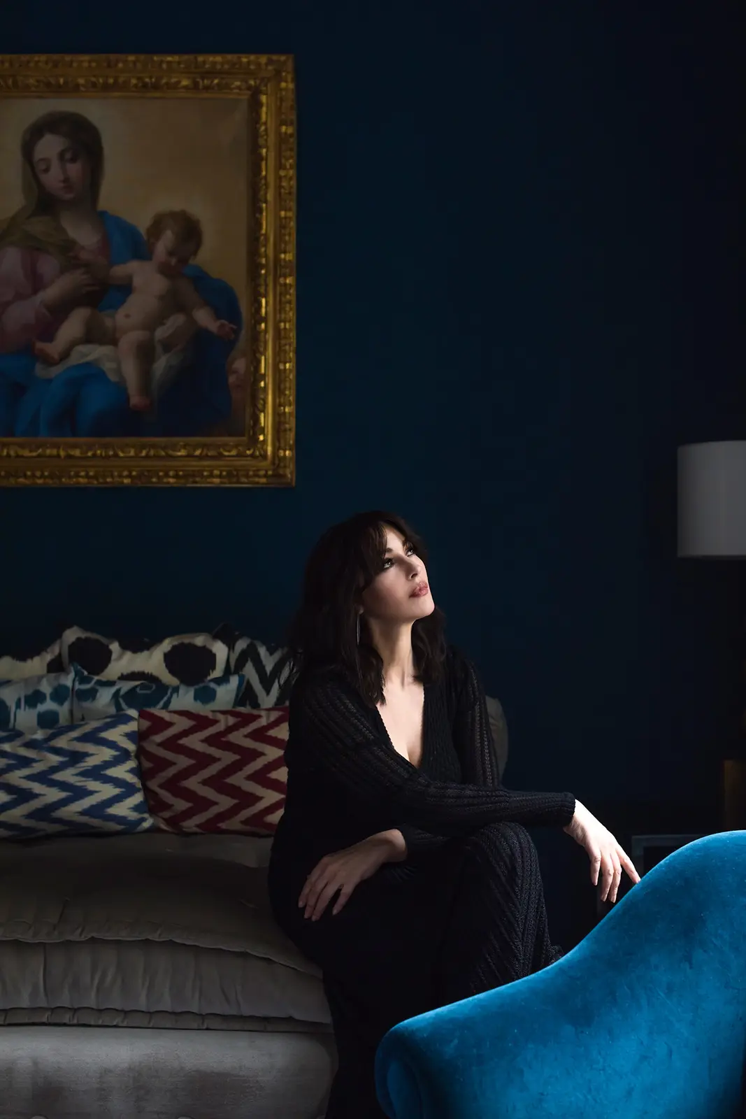 A woman in a black dress sits on a couch looking upward, with a classical painting of the Virgin Mary holding the baby Jesus…
