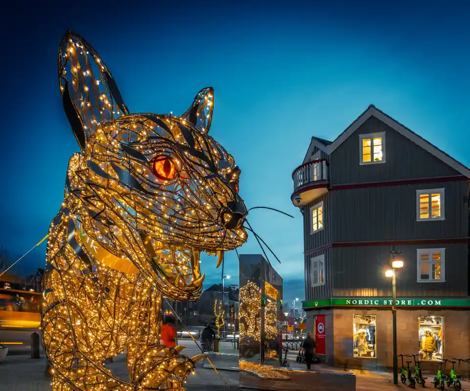 A large, illuminated wireframe sculpture of a cat with glowing orange eyes and sharp teeth stands in a city square at dusk…