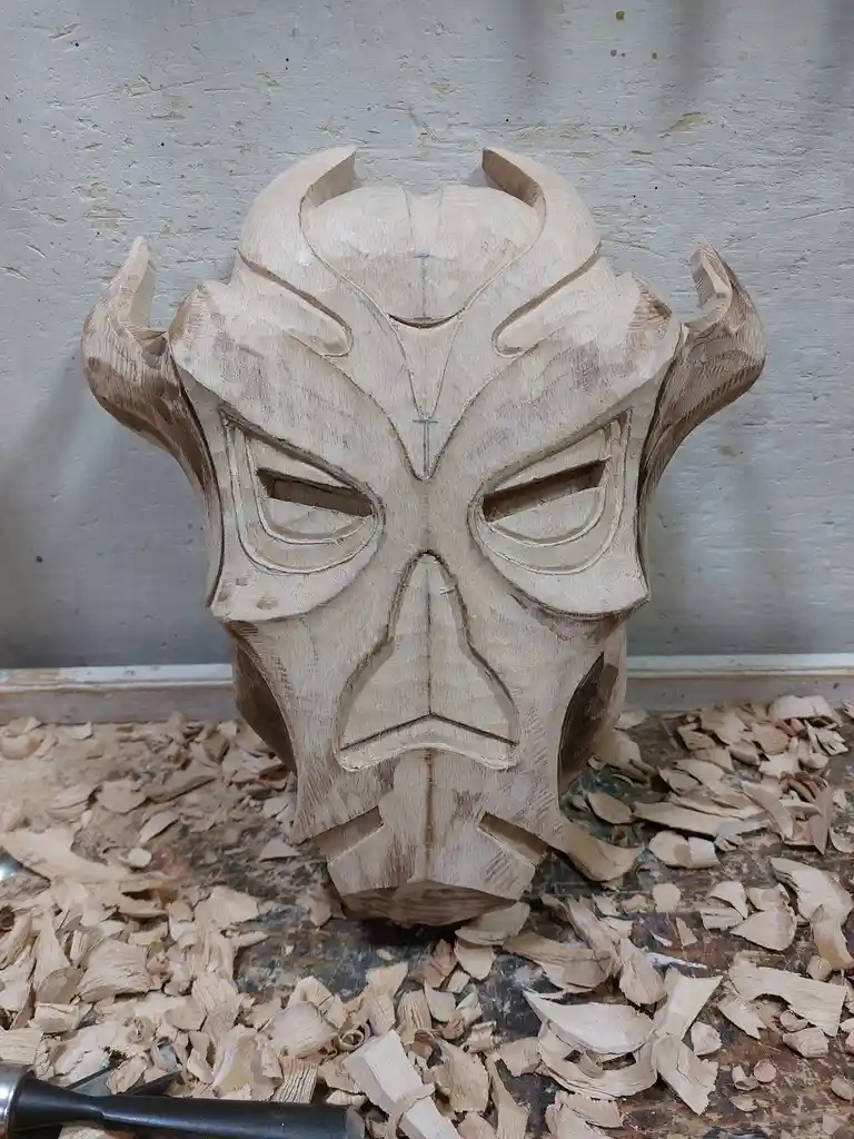 A wooden mask with carved horns and an angry expression sits on a workbench covered in wood shavings, with a chisel visible…