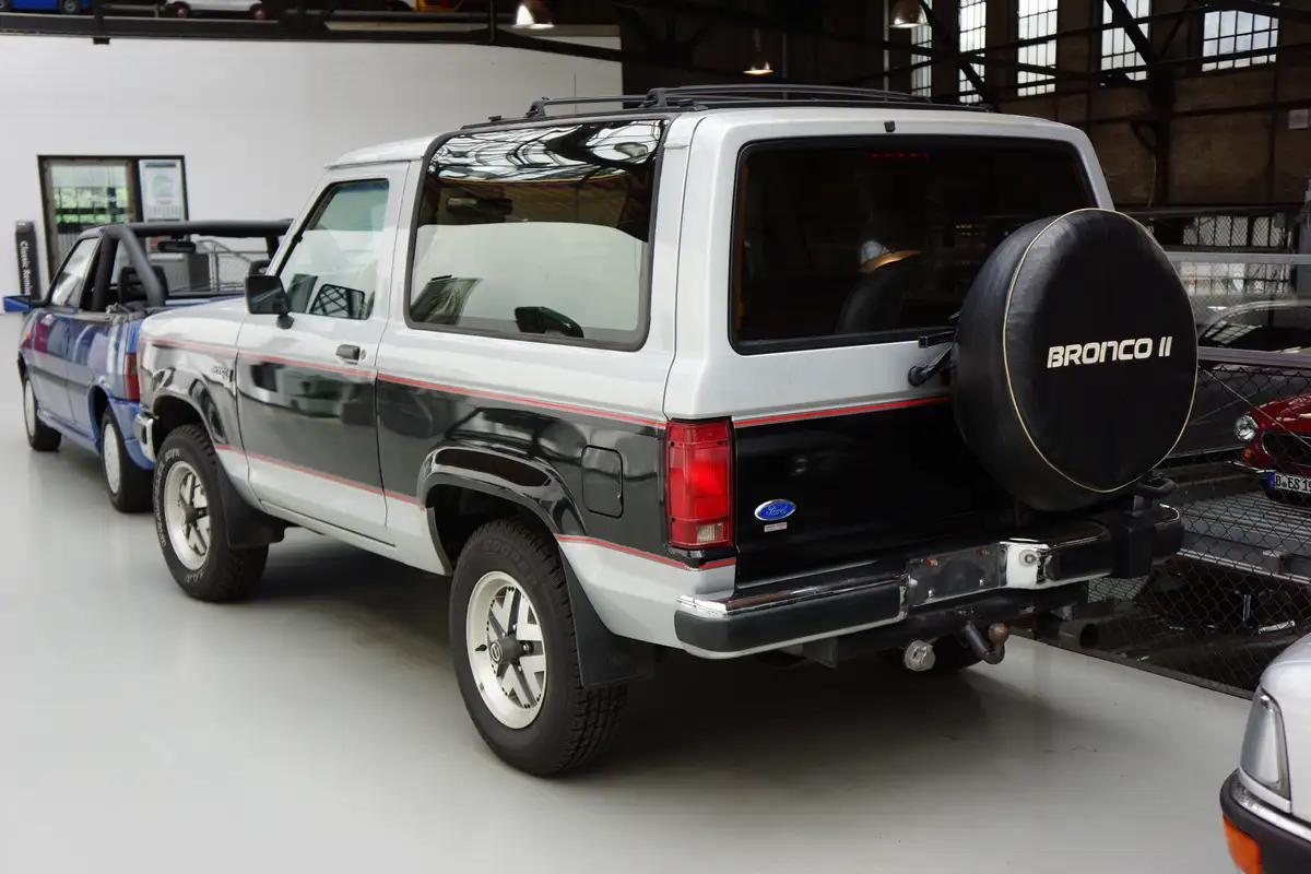 A white and black Ford Bronco II is shown from the rear in a car museum, featuring a spare tire cover with 'BRONCO II'…