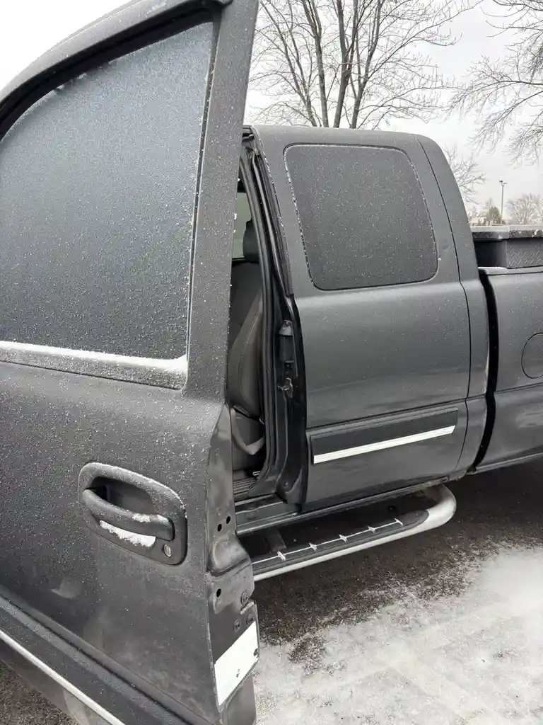 A close-up photo shows the open driver's side door of a dark gray pickup truck, with snow dusting the door, window, handle…