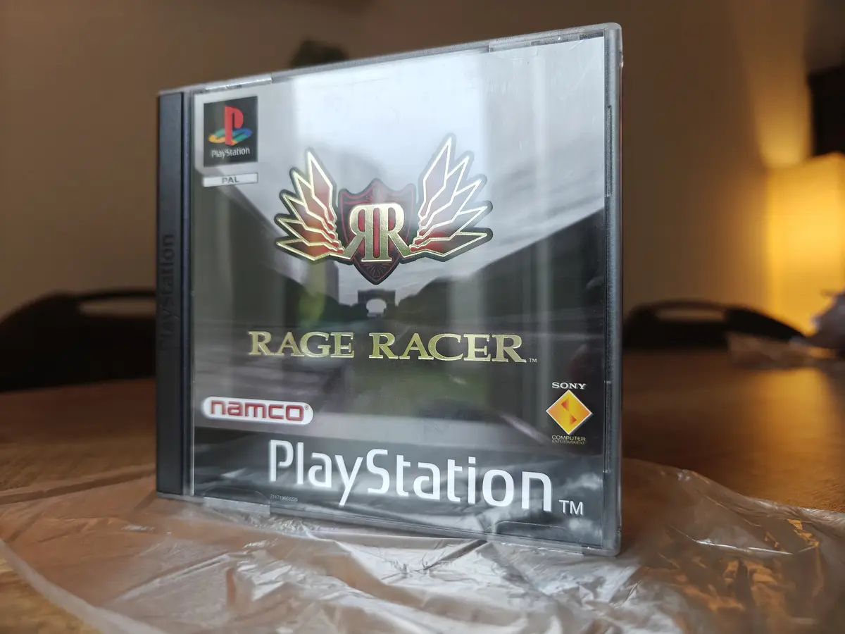 A physical PlayStation 1 game case for 'Rage Racer' by Namco, standing upright on a crinkled plastic surface. The cover art…