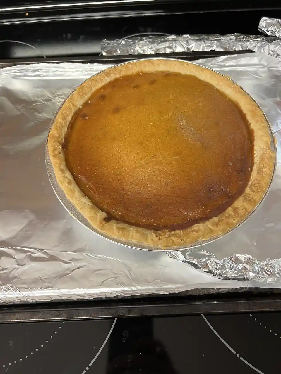 A freshly baked pumpkin pie sits in a metal pie tin on a baking sheet lined with aluminum foil, resting on a black stovetop…