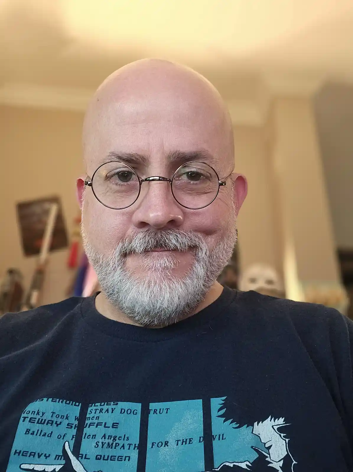 A bald man with a gray beard and round glasses smiles at the camera while wearing a dark t-shirt with a graphic of…