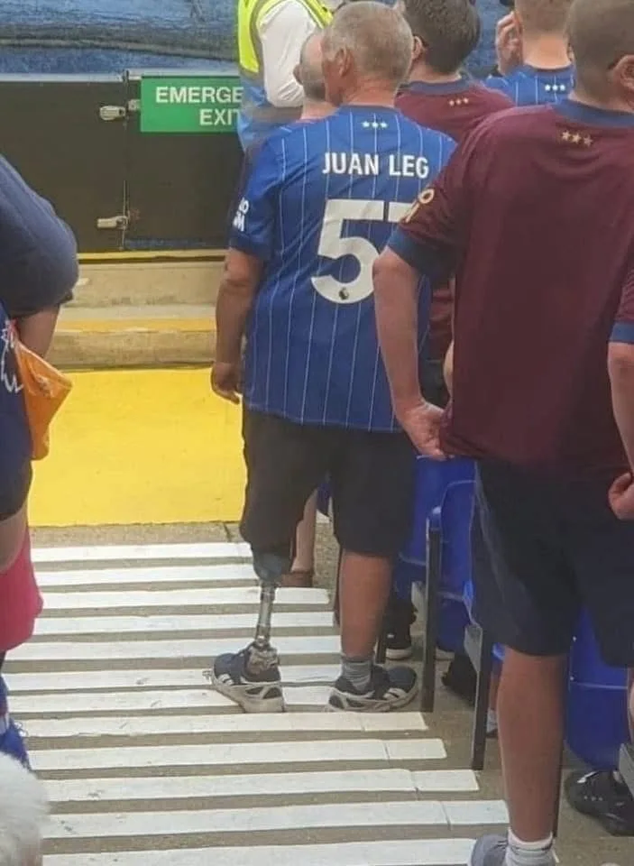 A man with a prosthetic leg stands on a zebra crossing, wearing a blue football jersey with 'JUAN LEG' and the number 57 on…