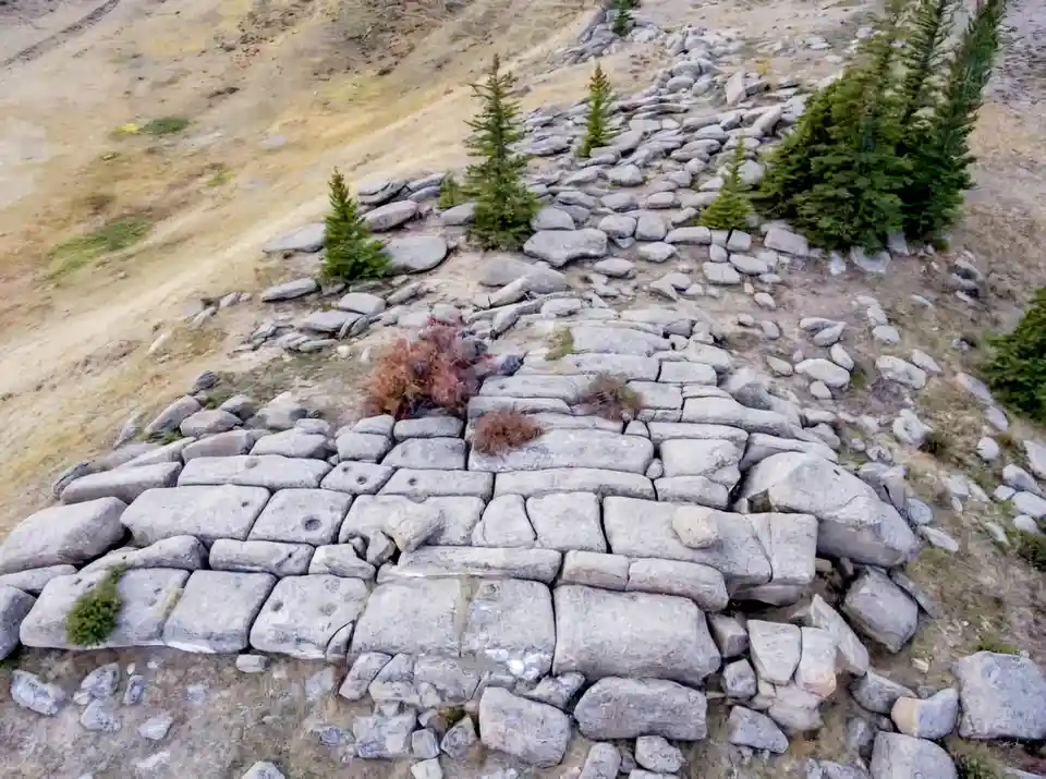 An aerial view shows a large, irregular stone wall built across a rocky, sparse hillside with scattered evergreen trees. The…