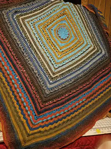 A close-up photo of a colorful, hand-crocheted afghan with concentric square patterns radiating outward from a central…