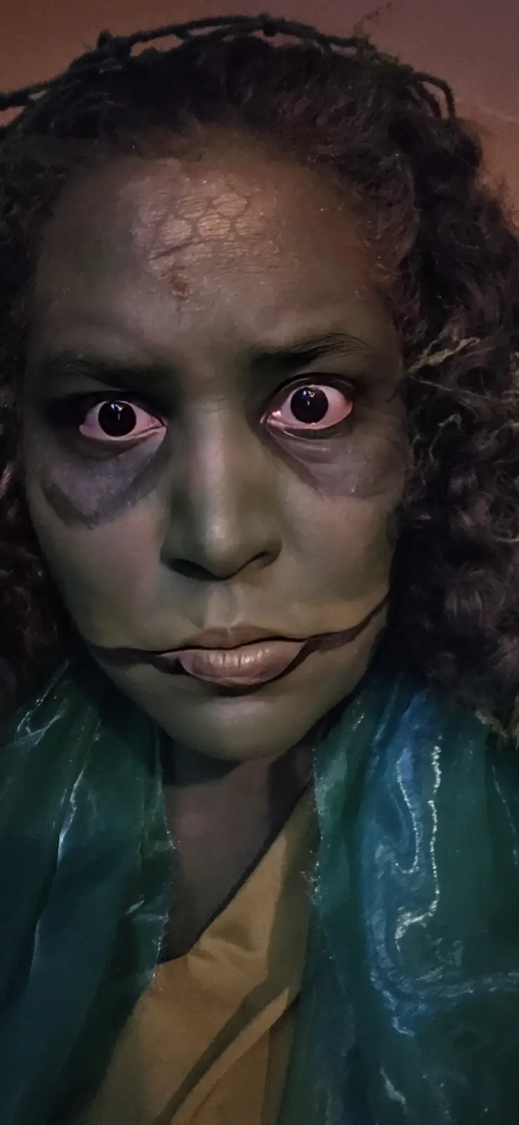 A person with dark curly hair wears dramatic green and black makeup resembling a horror character, with glowing red eyes and…