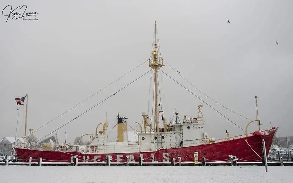 A red and white historic lightship named 'OVERFALLS' is docked in a snowy harbor under an overcast sky, with snow covering…