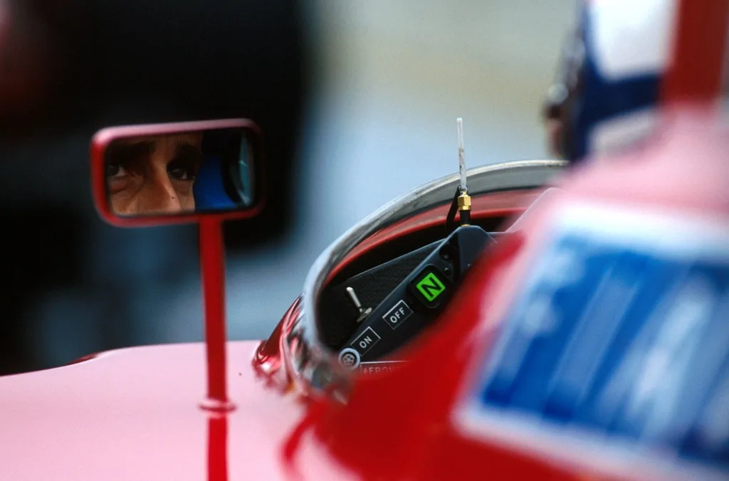 A close-up shot of a red Formula 1 car's side mirror, reflecting the intense gaze of the driver. The car's helmet visor is…
