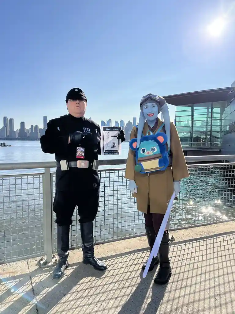 Two people in Star Wars cosplay stand on a waterfront promenade with a city skyline behind them. The person on the left…