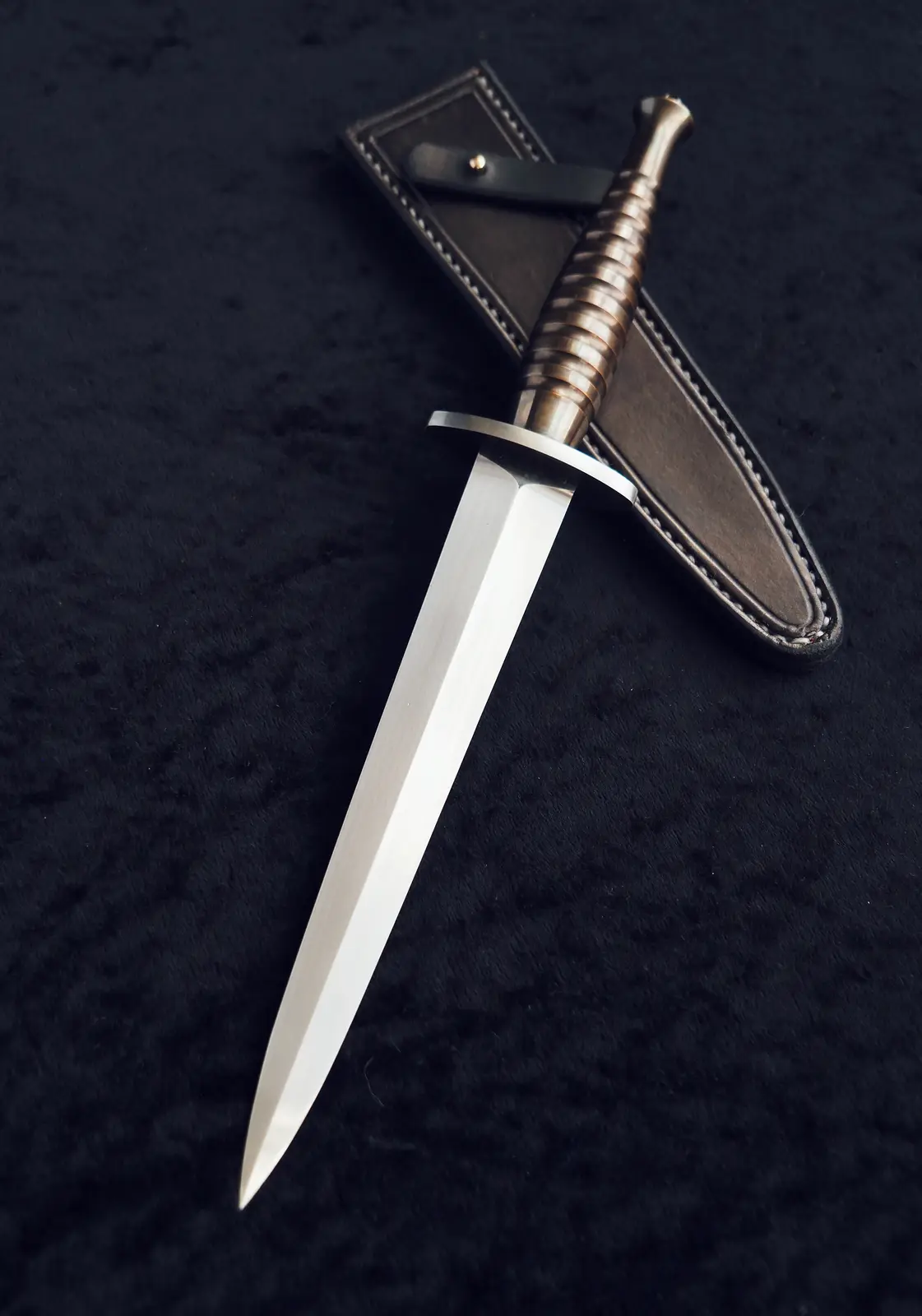 A close-up photograph shows a shiny, silver-bladed dagger with a brass-colored, ribbed handle resting diagonally on a dark…
