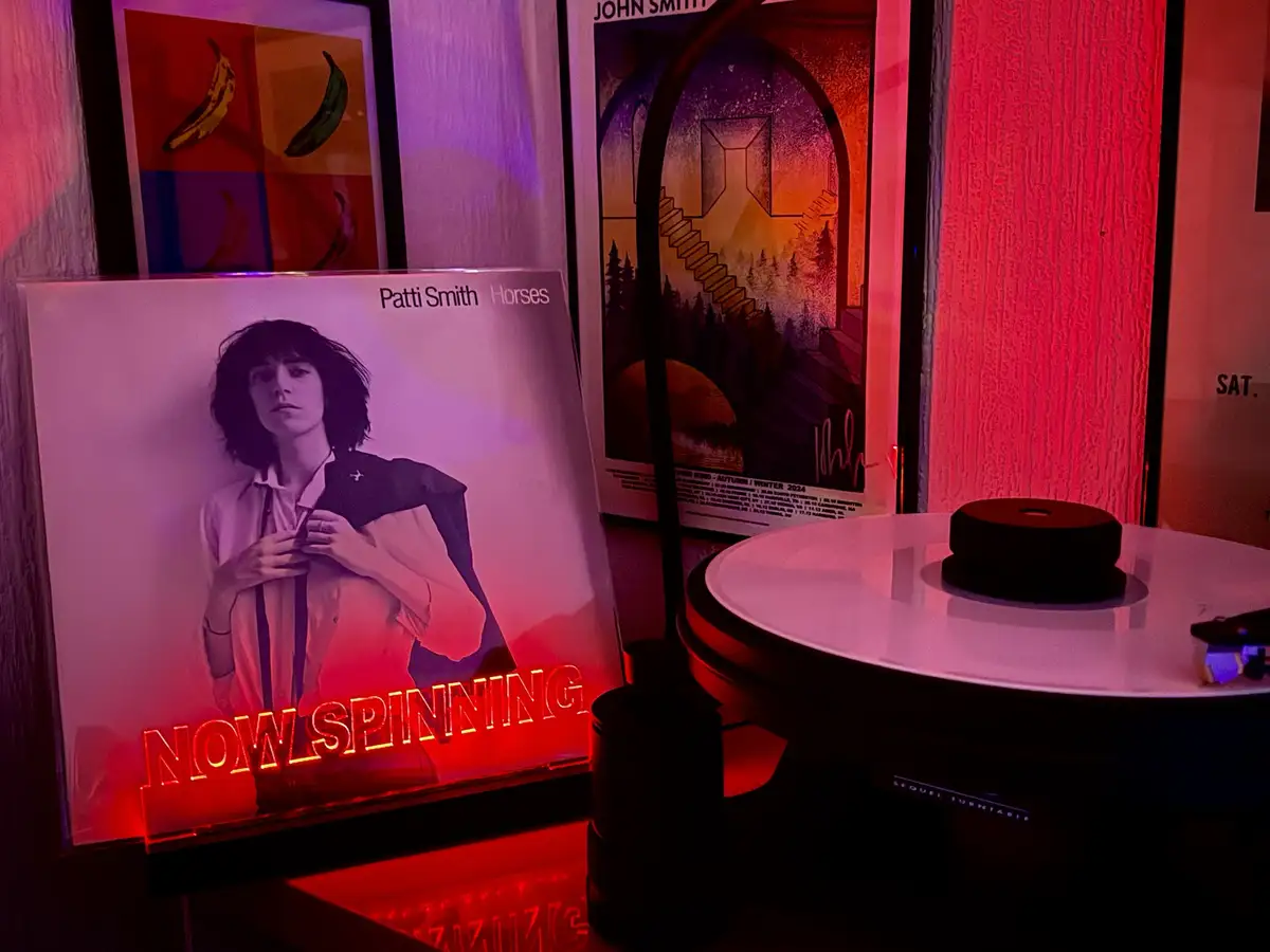 A vinyl record player sits next to a Patti Smith 'Horses' album cover, which has a glowing red neon sign reading 'NOW…