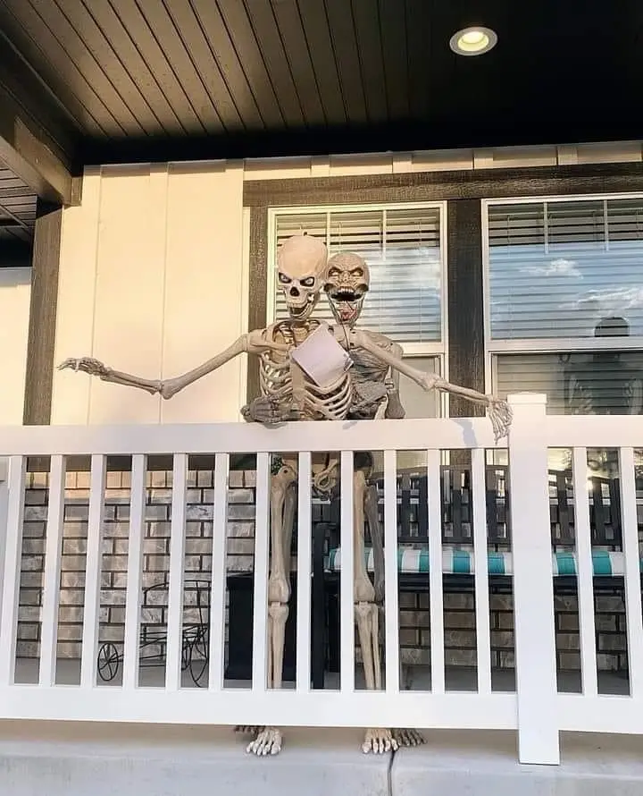 Two Halloween skeleton decorations stand on a porch behind a white railing, one holding a piece of paper. The skeleton on…