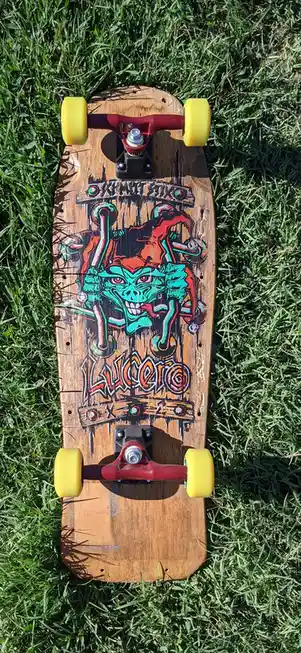 A longboard skateboard with a wooden deck, red trucks, and yellow wheels rests on green grass. The deck features a colorful…