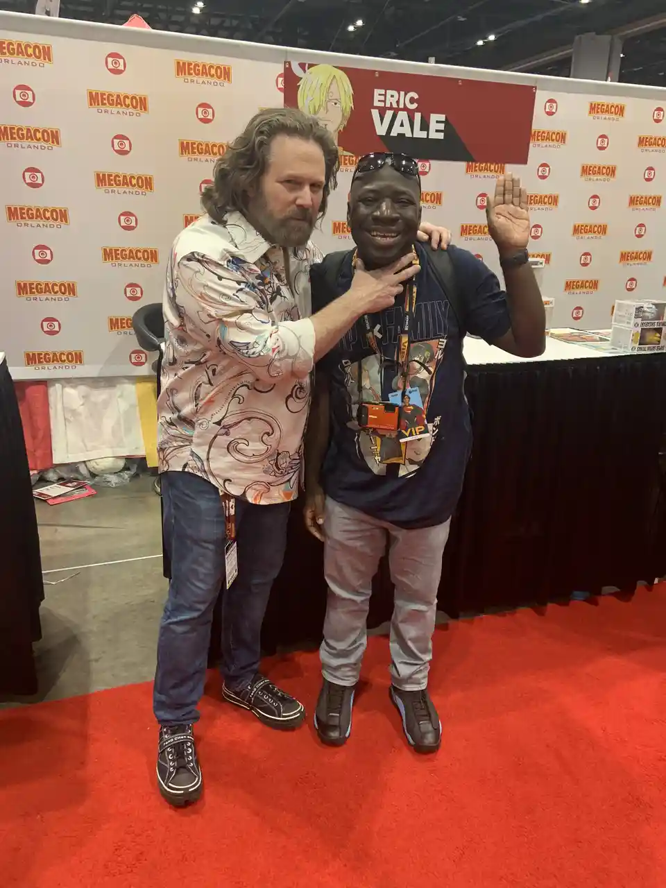 Two men pose for a photo on a red carpet at Megacon Orlando. The man on the left, with long hair and a patterned shirt, has…
