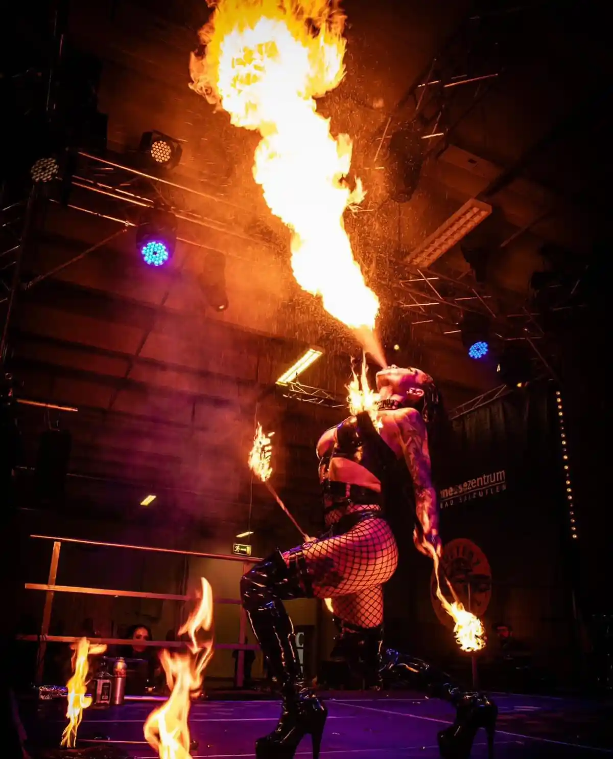 A fire dancer in a black outfit and fishnet stockings performs on stage, blowing a large stream of fire from her mouth while…