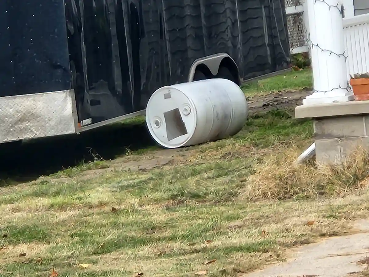 A white metal barrel lies on its side on grass next to a black trailer, with a square cutout on its end resembling a cat's…