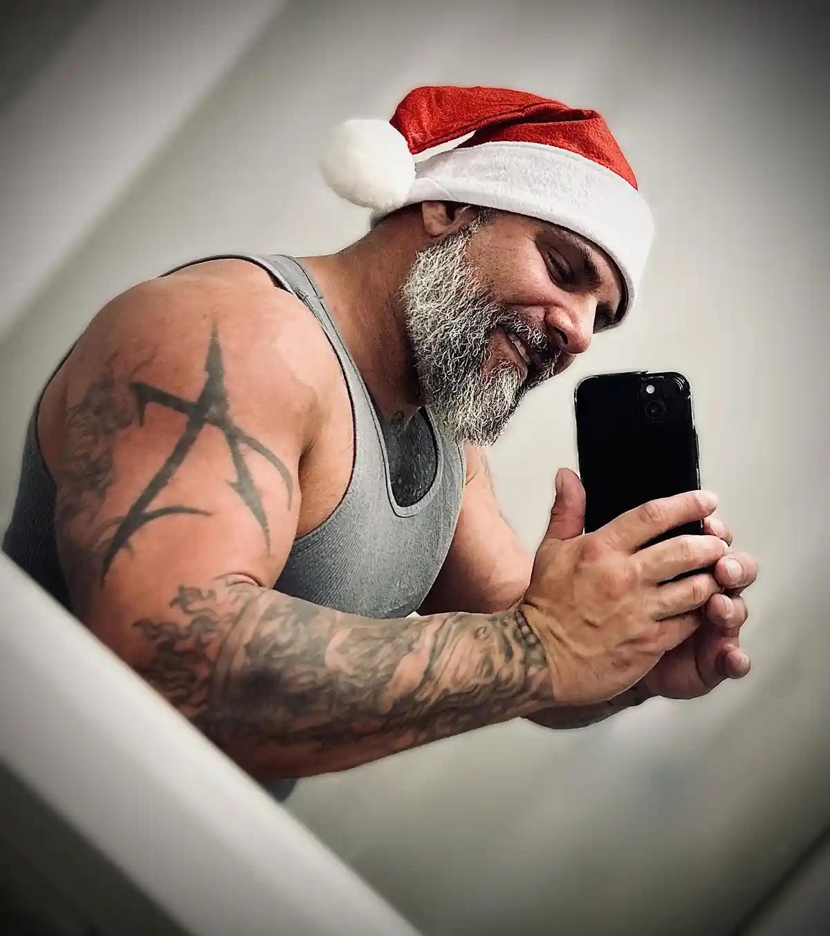 A muscular man with a grey beard and tattoos on his arms wears a red and white Santa hat while smiling and looking at his…