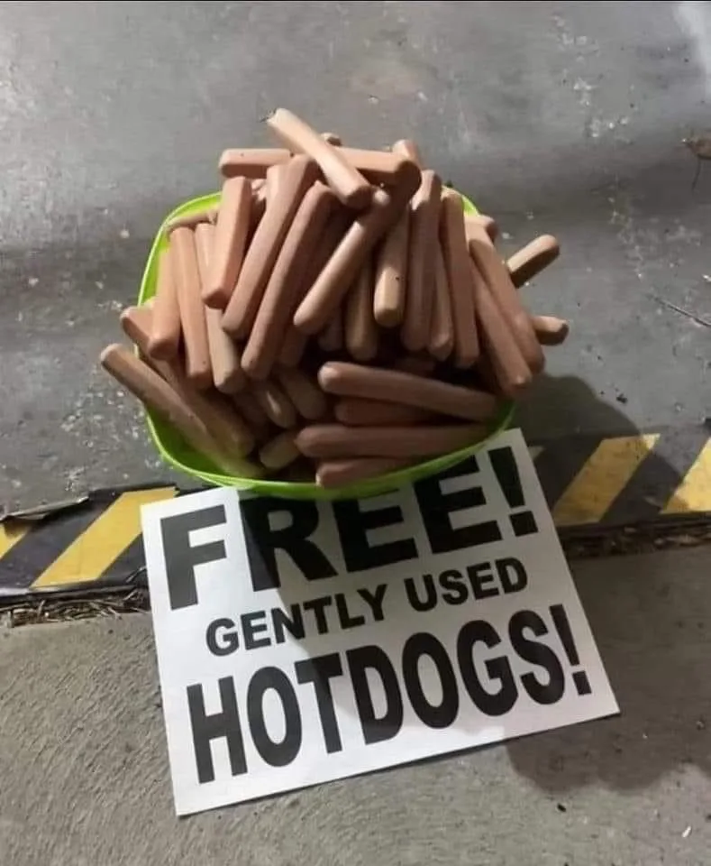 A green bowl overflowing with raw, uncooked hot dogs sits on a concrete floor next to a sign that reads 'FREE! GENTLY USED…