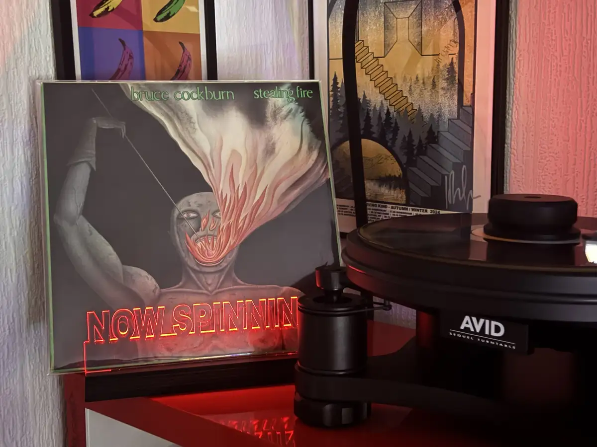A vinyl record album titled 'Stealing Fire' by Bruce Cockburn is standing upright next to an AVID Sequel turntable. The…