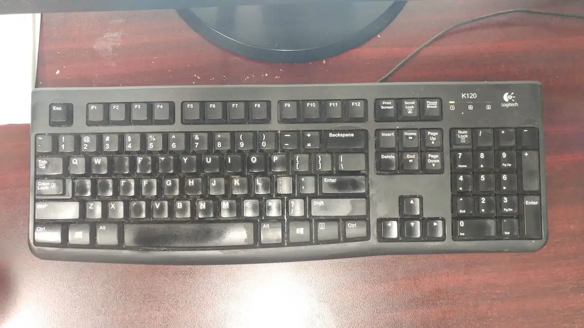 A black Logitech K120 keyboard sits on a wooden desk, showing significant wear and dirt on its keys, with a monitor base…