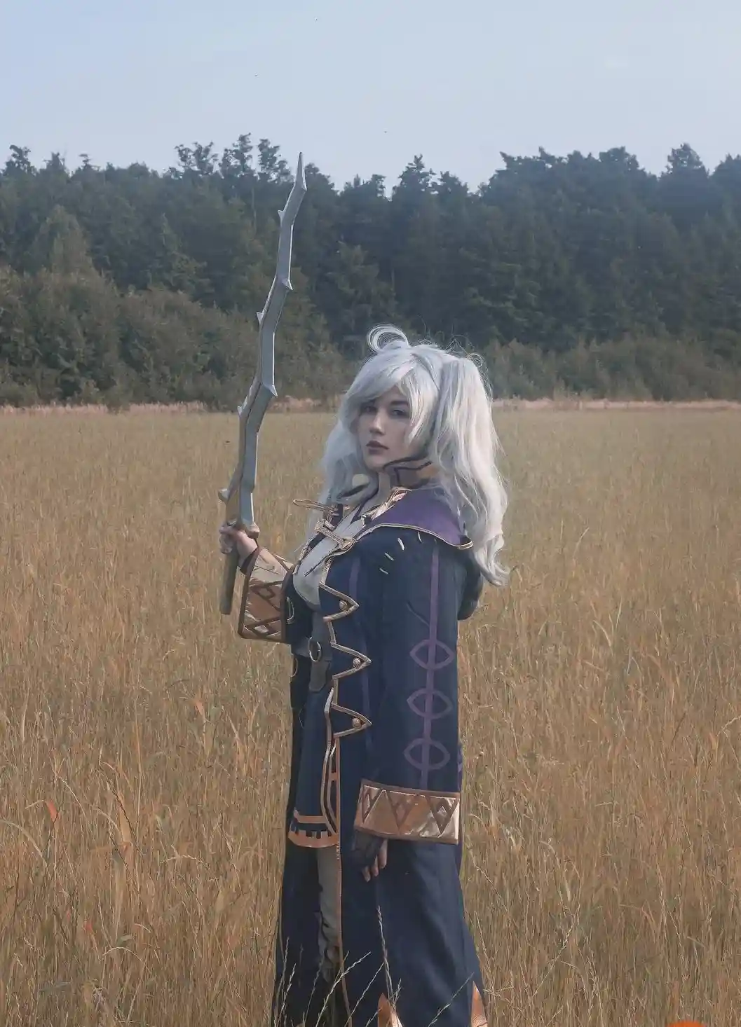 A person with long silver hair and a serious expression stands in a field of tall, dry grass, holding a large, jagged sword…