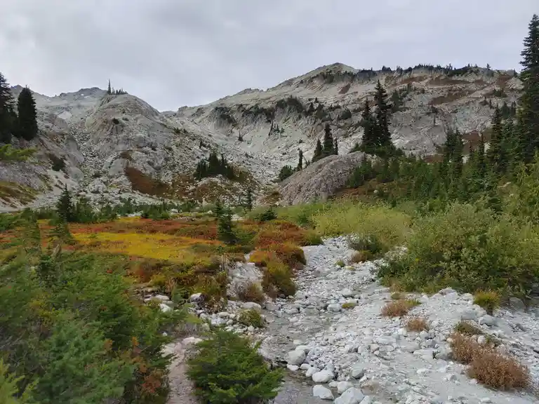 A scenic mountain landscape with rocky terrain, evergreen trees, and colorful autumn foliage under an overcast sky. A gravel…
