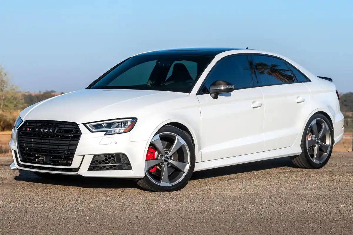 A white Audi S3 sedan with black roof and red brake calipers is parked on gravel under a clear blue sky. The overlaid text…