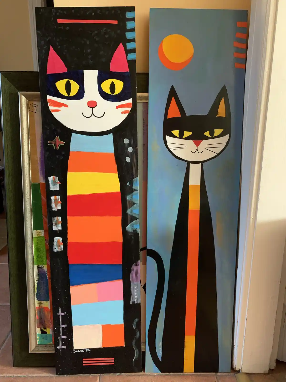 Two tall, stylized cat paintings stand side by side; the left cat wears a colorful striped scarf against a black background…