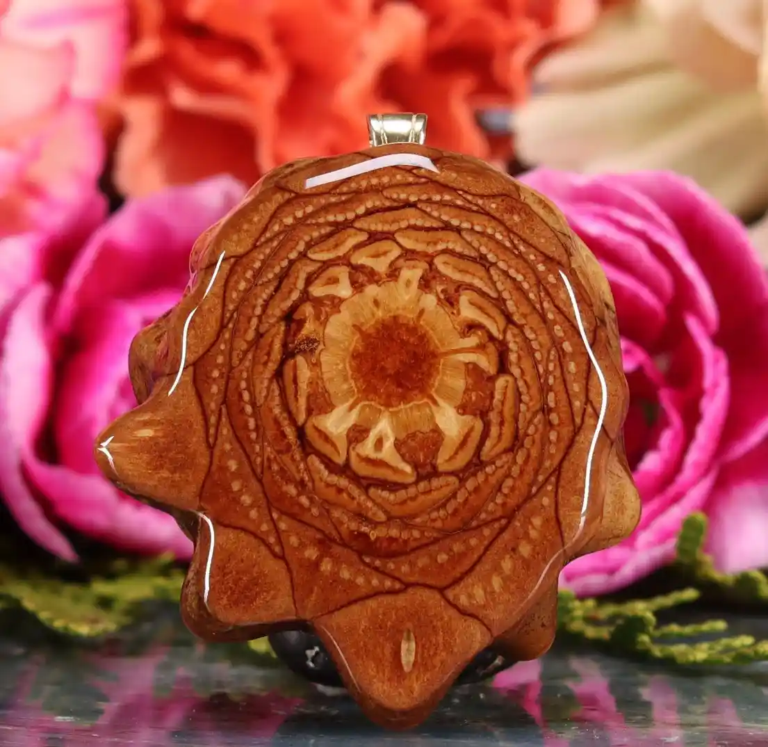 a polished wooden pendant shaped like a rose with intricate carved patterns, displayed against a backdrop of pink and orange…