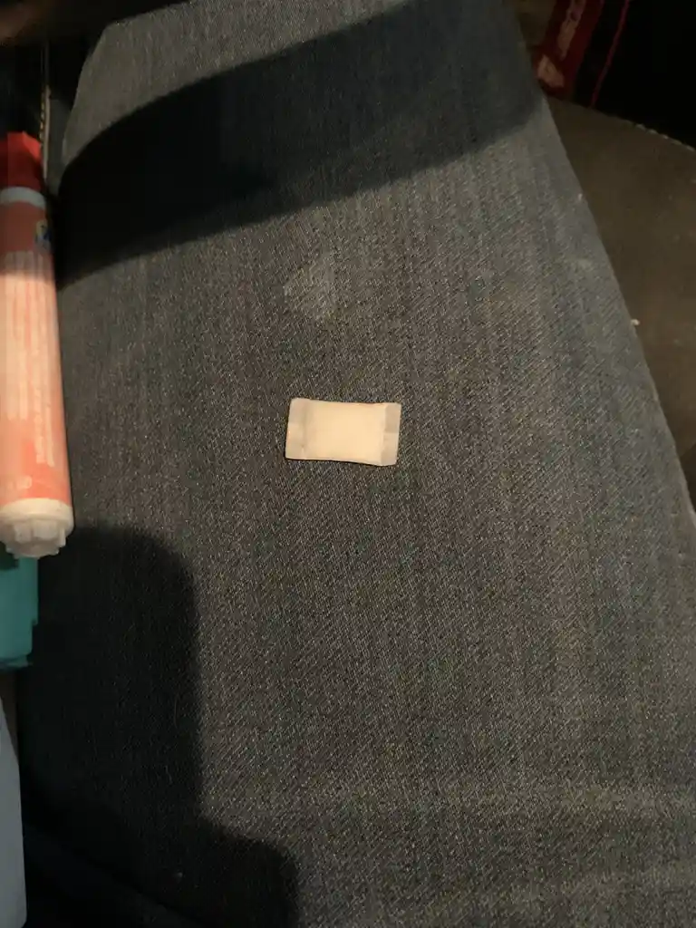 A small, rectangular adhesive bandage lies centered on a dark gray fabric surface, likely a couch or chair. To the left, a…