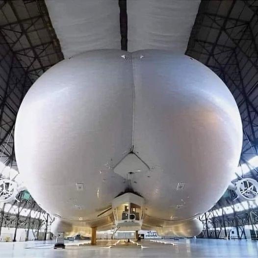 a large, white, spherical object resembling a human buttocks is shown from a low angle inside a hangar, with the overlaid…
