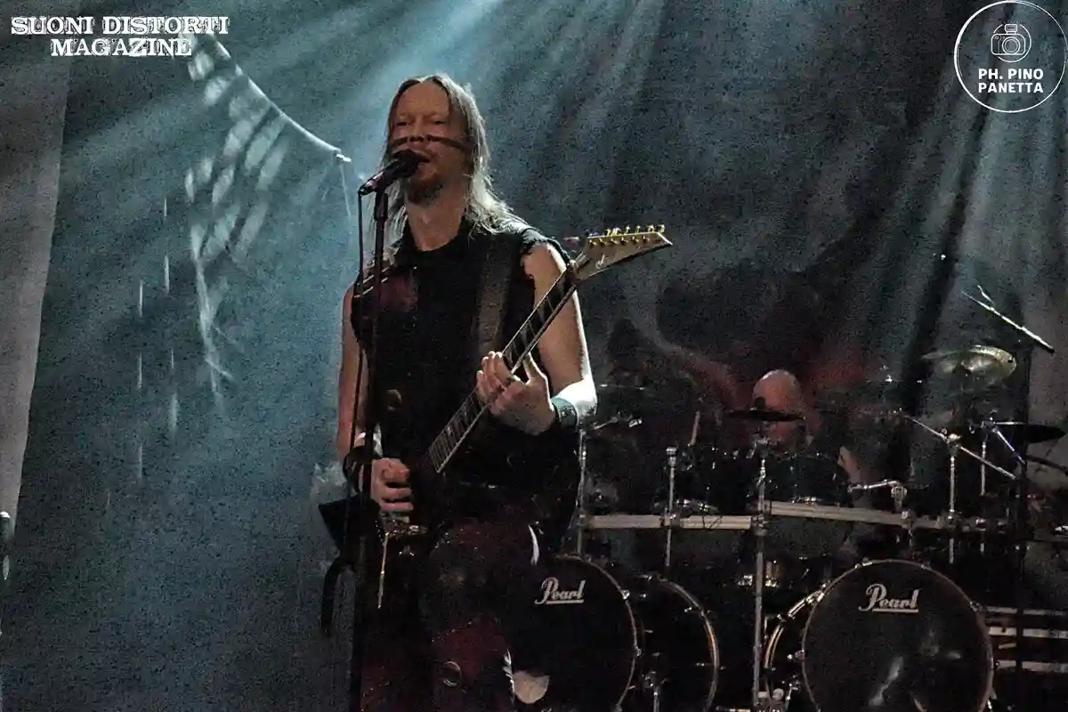A musician with long hair, wearing a black sleeveless top and holding an electric guitar, sings into a microphone on a dimly…