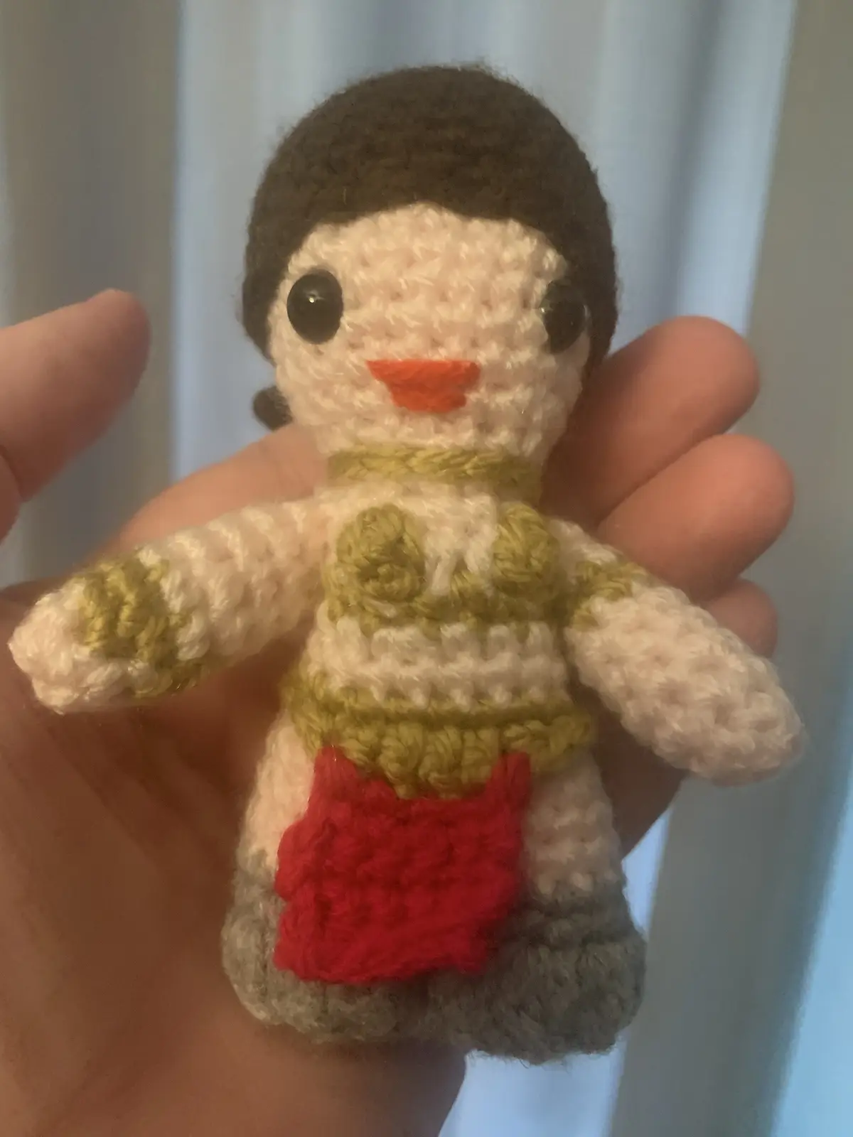 A hand holds a small, crocheted amigurumi doll with brown hair, black button eyes, an orange mouth, and a green and white…