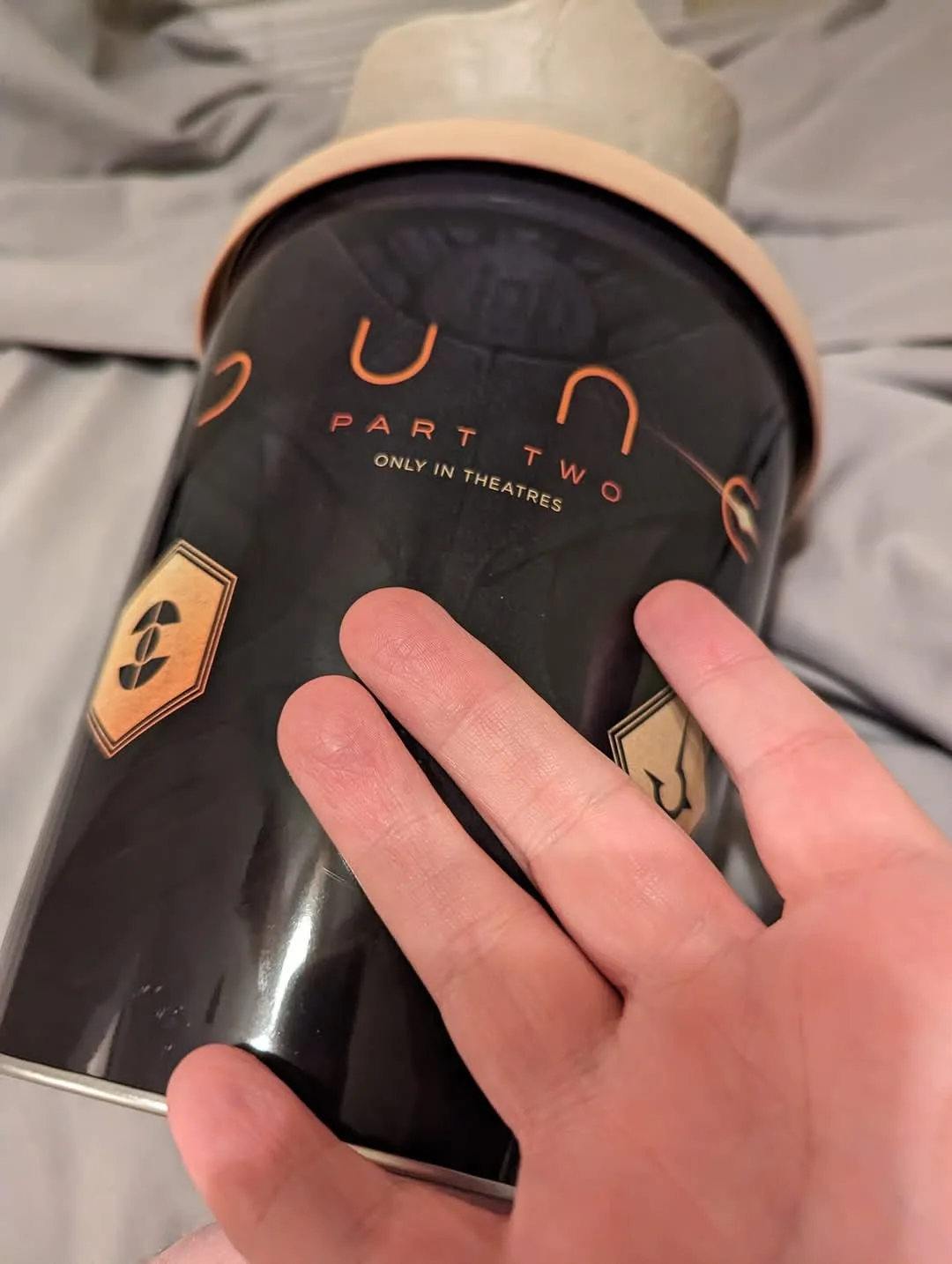 A hand holds a black cylindrical Dune Part Two movie prop or collectible, which has orange text reading 'DUNE PART TWO ONLY…