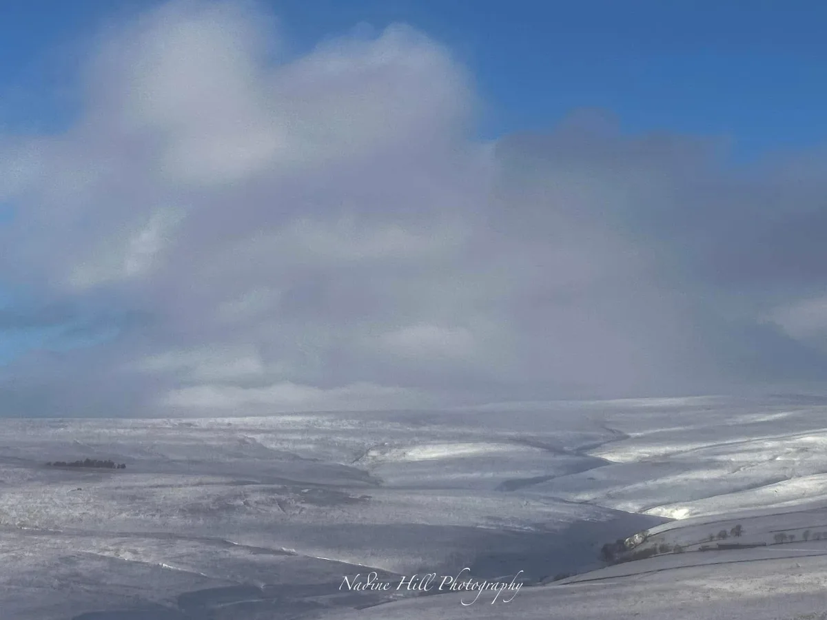 A wide, snowy landscape under a partly cloudy blue sky, with rolling hills covered in white snow and sparse trees in the…