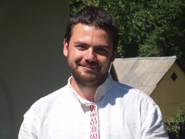 A man with short dark hair and a beard, wearing a white traditional shirt with red embroidery, smiles slightly while…